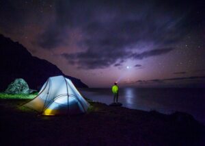 Night time image of tent next to body of water and camper looking out at start sky.